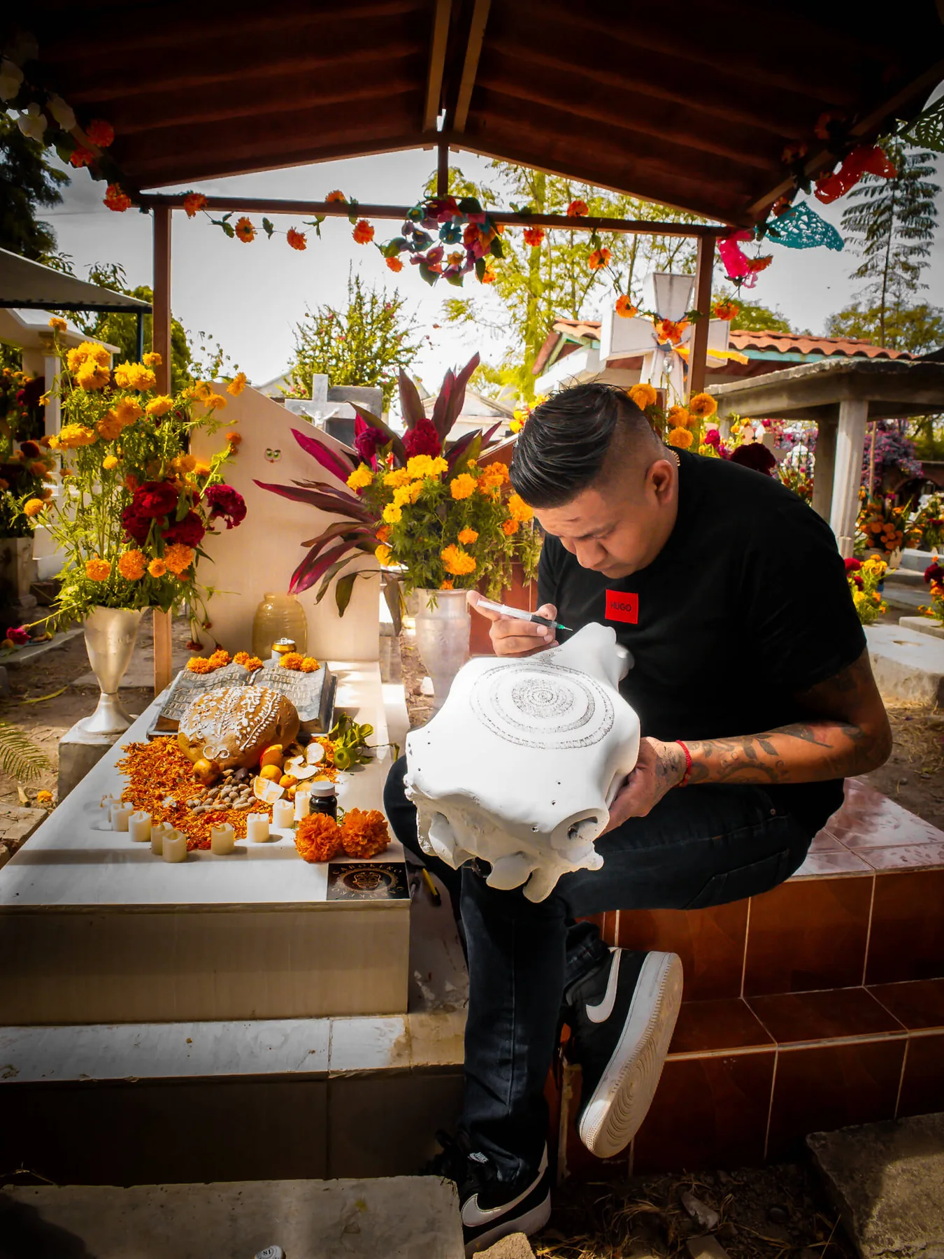 Ricardo painting a white skull beside a family ofrenda filled with flowers, bread and candles